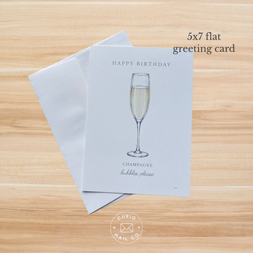 A set of 5x7 flat greeting cards and envelopes on a light wood background. The front card is light gray with a minimalist illustration of a champagne flute and the text 'HAPPY BIRTHDAY' and 'CHAMPAGNE bubbles, please'.