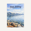 Scenic Lake Tahoe birthday card with blue water, rocky shoreline, mountains, and Happy Birthday text