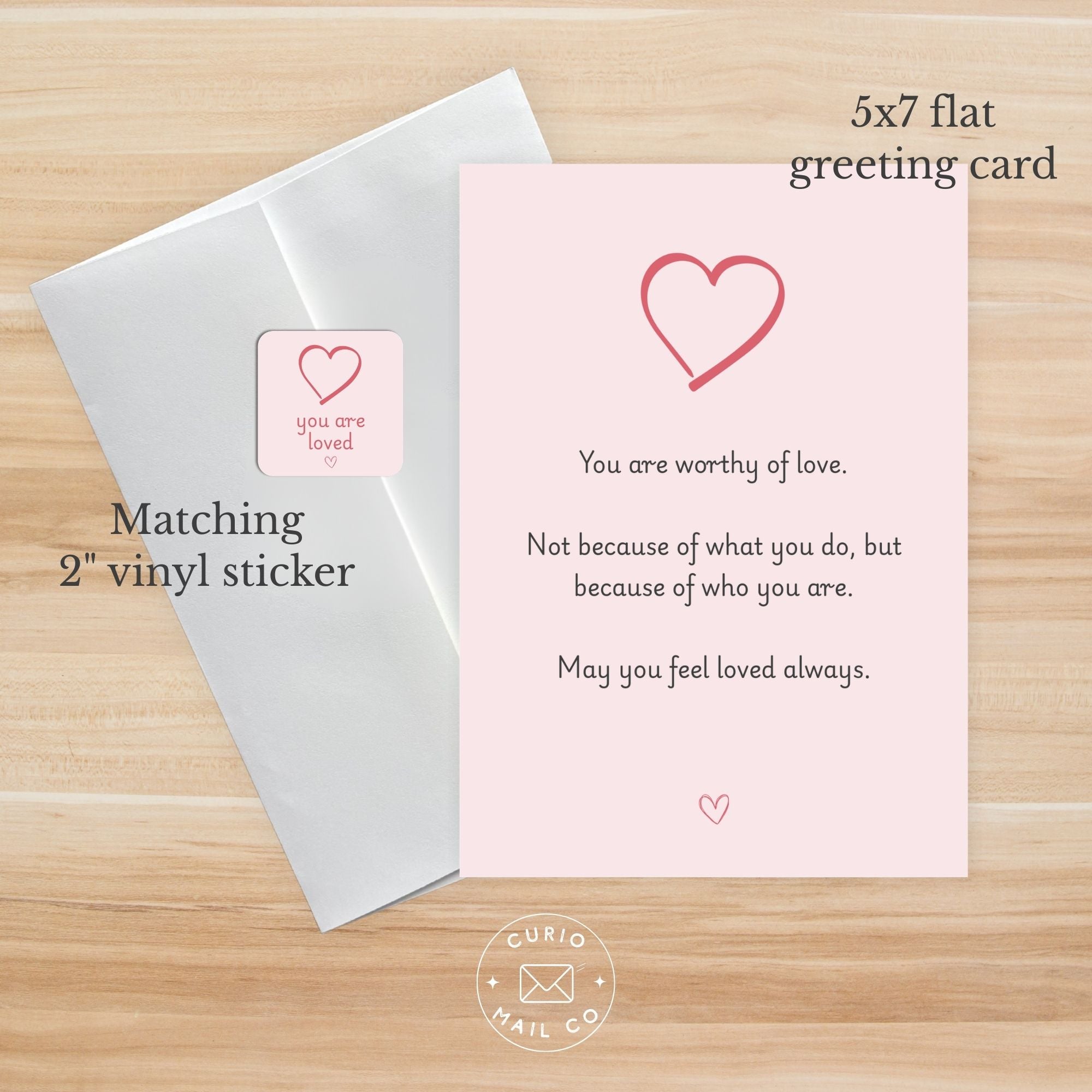 Pink greeting card with heart design and text, matching white envelope, and pink vinyl sticker on a wooden surface.