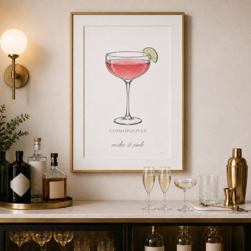 A framed art print featuring a pink cocktail in a coupe glass with a lime wedge, labeled 'COSMOPOLITAN make it pink', displayed on a beige wall above a bar counter with bottles, glasses, and a gold shaker.