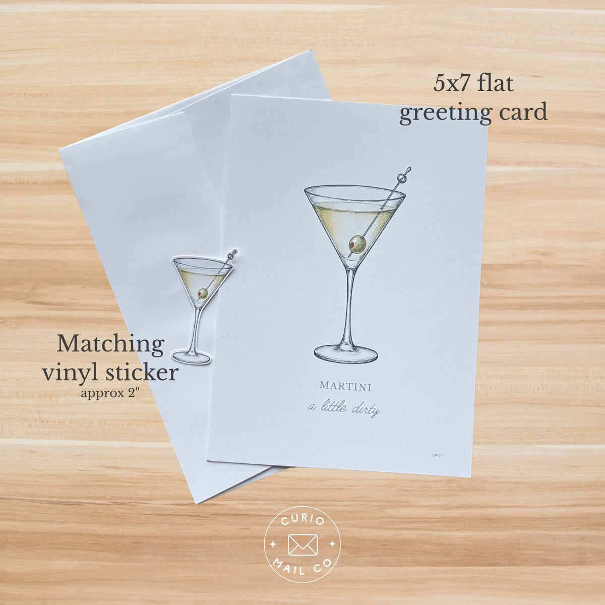 A 5x7 flat greeting card and matching vinyl sticker are shown on a light wood background. The card features a minimalist illustration of a martini glass with an olive, and the sticker is slightly smaller with the text 'Matching vinyl sticker approx 2"'.